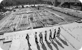 A group of construction workers in hard hats stand on-site, overlooking a large foundation with rebar laid out for a building project.