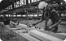 A construction worker in safety gear uses tools on a wooden beam at a building site, surrounded by equipment and materials.