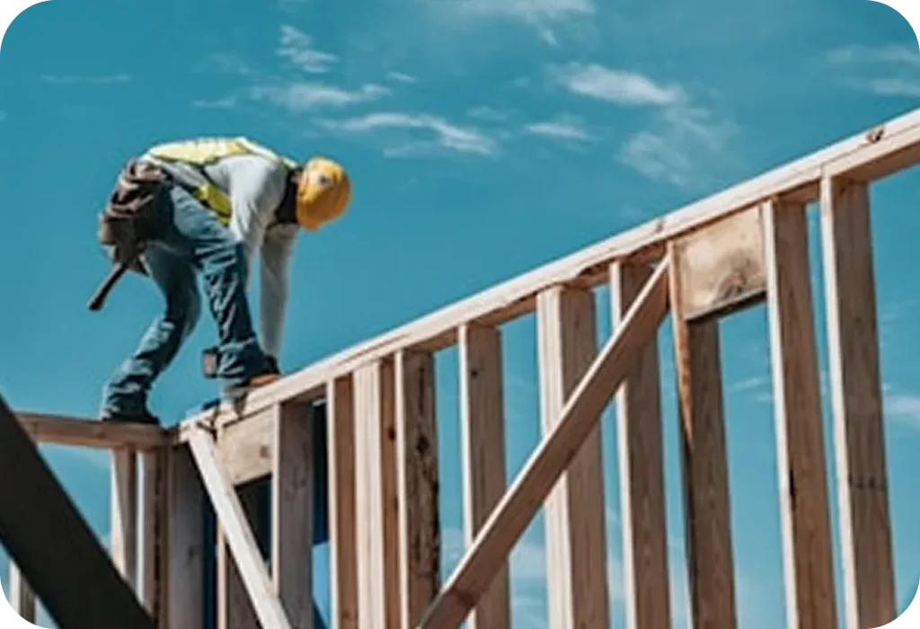 A construction worker in safety gear and helmet works on framing a wooden structure under a clear blue sky.