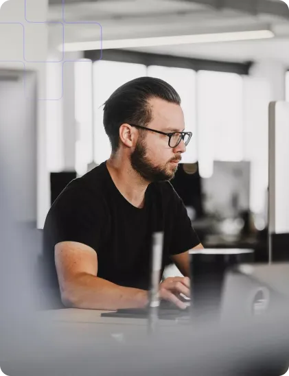 A man with glasses and a beard, dressed in a black t-shirt, works intently on a computer in a bright office setting.