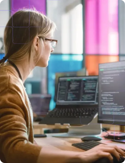 A woman works at a desk with dual monitors, focused on coding and technical tasks in a modern office setting.