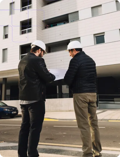 Two construction professionals wearing hard hats review plans outside a modern building site.