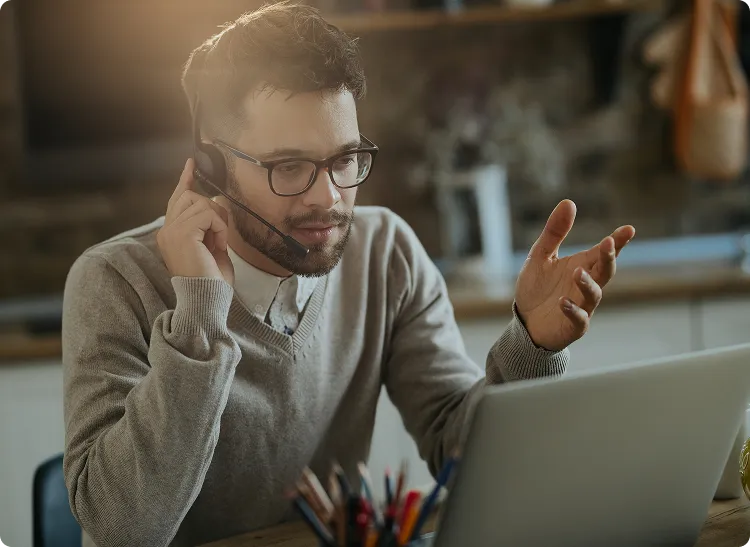 A man wearing glasses and a headset is speaking while working on a laptop, likely providing online support or training.