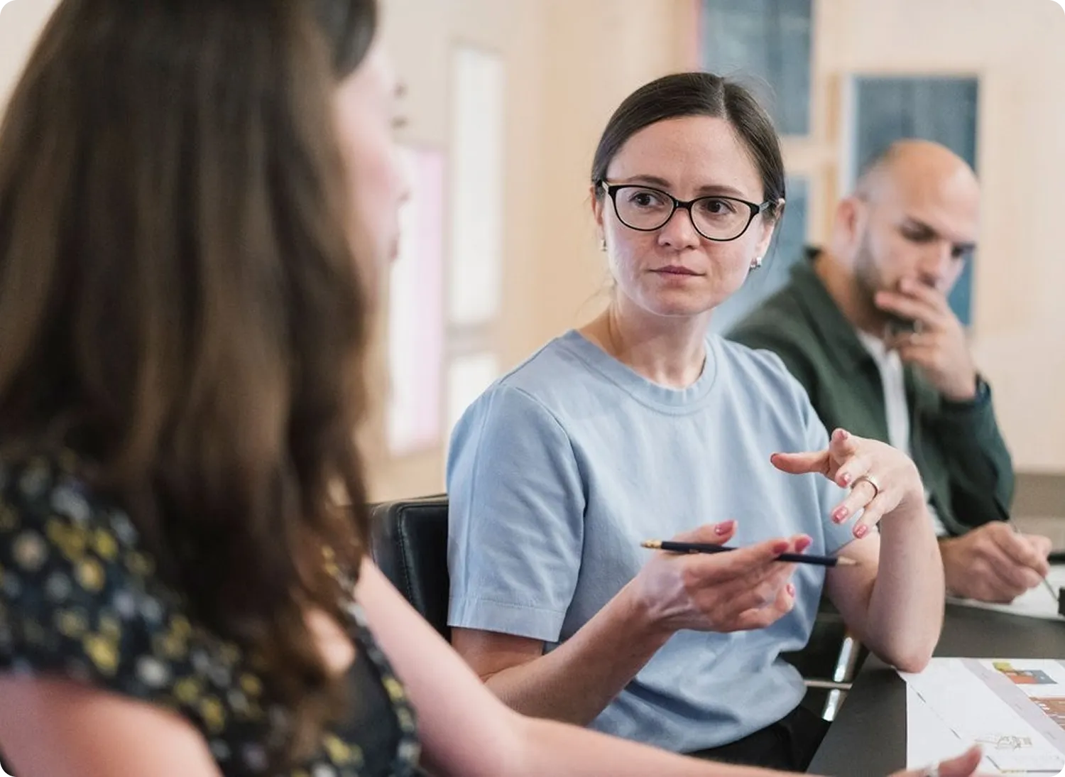 A woman in glasses and a light blue shirt is speaking while holding a pen, engaging with another woman in the foreground.