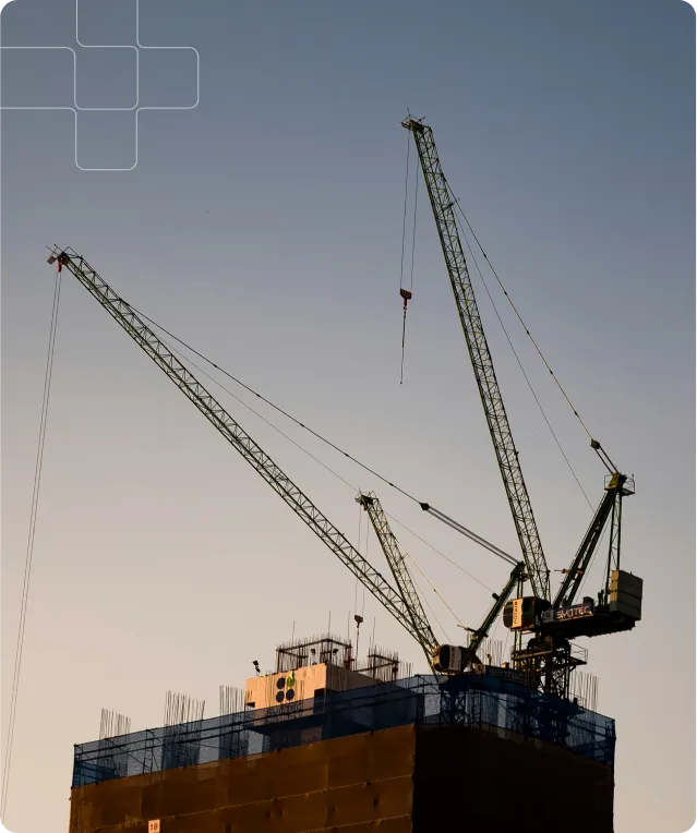 Two cranes sit atop a building under construction. The building is covered in blue netting and the cranes are green. The sky is a light blue.