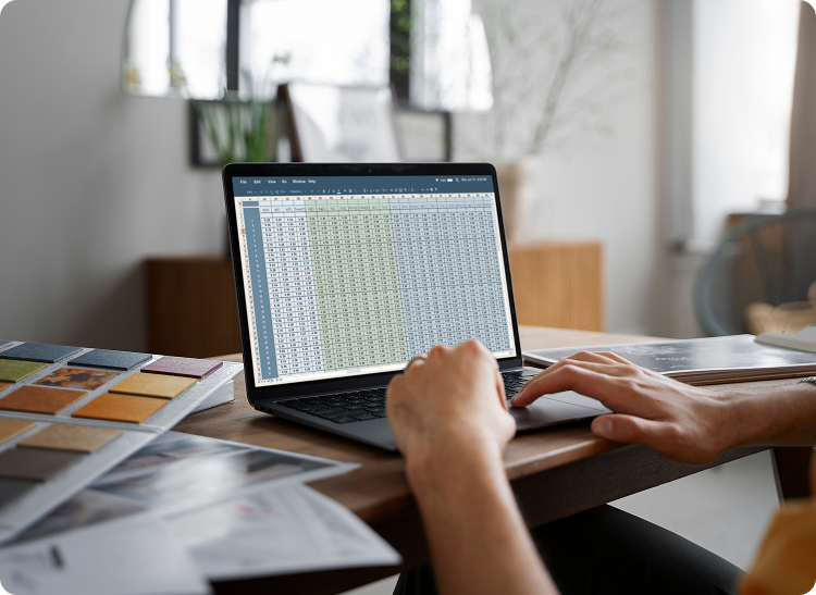 Person working on a laptop displaying a spreadsheet, with documents spread on the desk.
